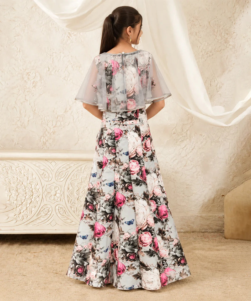 A woman viewed from behind wearing a long floral printed gown with sheer flutter sleeves and a flared skirt, standing indoors against a cream textured wall and draped fabric.