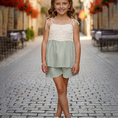 A young girl stands on a cobblestone street wearing a two-piece summer outfit consisting of a light sage green and cream lace top with matching shorts.
