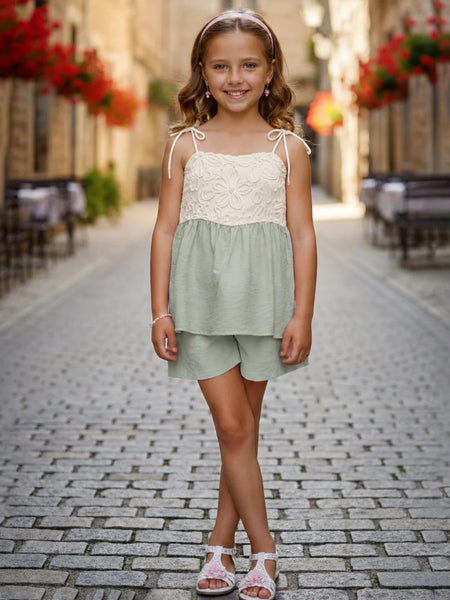 A young girl stands on a cobblestone street wearing a two-piece summer outfit consisting of a light sage green and cream lace top with matching shorts.
