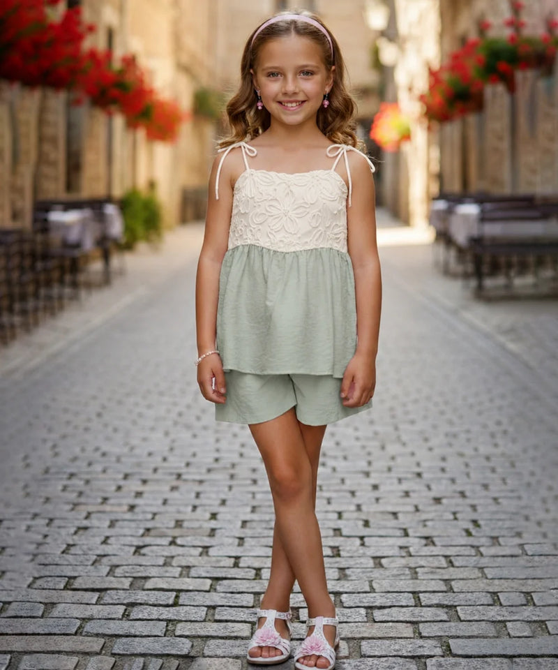 A young girl stands on a cobblestone street wearing a two-piece summer outfit consisting of a light sage green and cream lace top with matching shorts.
