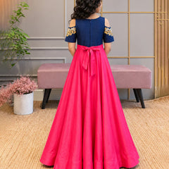 A young girl is shown from behind wearing a formal A-line dress with a navy blue bodice, cold-shoulder sleeves with gold embroidery, and a bright pink skirt with a matching pink bow at the waist. She is standing in an indoor setting with a bench and plants in the background.