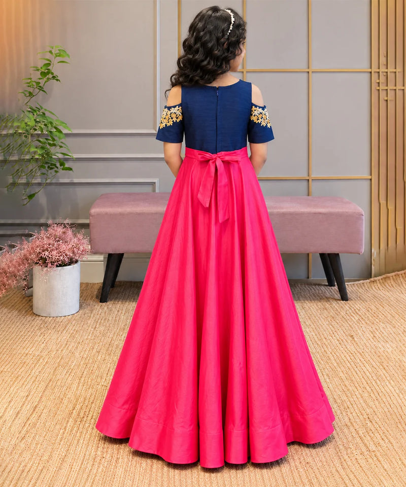 A young girl is shown from behind wearing a formal A-line dress with a navy blue bodice, cold-shoulder sleeves with gold embroidery, and a bright pink skirt with a matching pink bow at the waist. She is standing in an indoor setting with a bench and plants in the background.