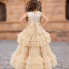 A young girl viewed from behind wearing a sleeveless champagne beige ball gown with a heavily embellished beaded bodice and a tiered tulle skirt, standing outdoors in front of a warm stone building.