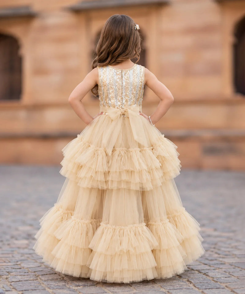 A young girl viewed from behind wearing a sleeveless champagne beige ball gown with a heavily embellished beaded bodice and a tiered tulle skirt, standing outdoors in front of a warm stone building.