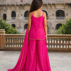 A woman stands with her back to the camera wearing a bright fuchsia pink traditional outfit with a matching dupatta, featuring gold border detailing, set against a historic stone courtyard with a palace facade in the background.