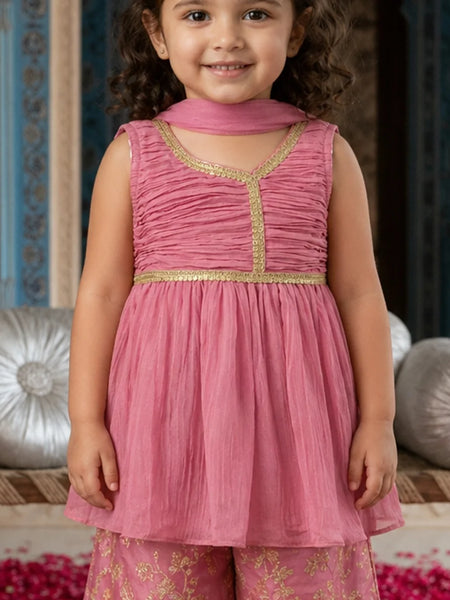 Young girl wearing a pink traditional outfit with gold details, standing against a decorative background.