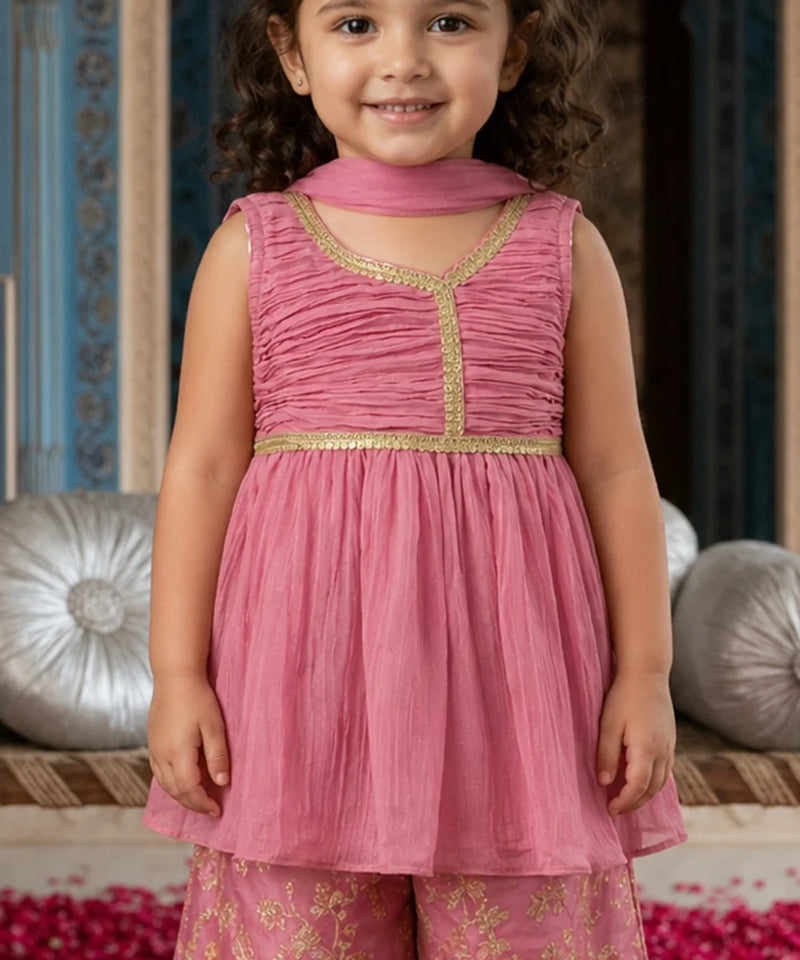 Young girl wearing a pink traditional outfit with gold details, standing against a decorative background.