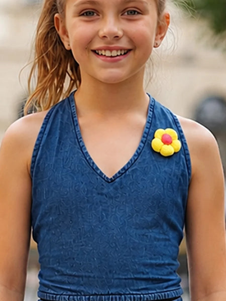 A smiling young girl wearing a blue denim-textured halter top with a yellow flower pin on the chest, photographed outdoors with a blurred background.