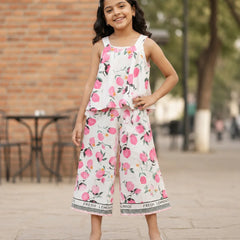 A young girl is shown from behind wearing a white sleeveless jumpsuit with a pink and green watercolor floral print. The jumpsuit features ruffled straps and a wide leg, with a black and white patterned hem that reads 'LEMONADE'.