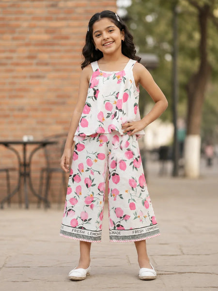 A young girl is shown from behind wearing a white sleeveless jumpsuit with a pink and green watercolor floral print. The jumpsuit features ruffled straps and a wide leg, with a black and white patterned hem that reads 'LEMONADE'.