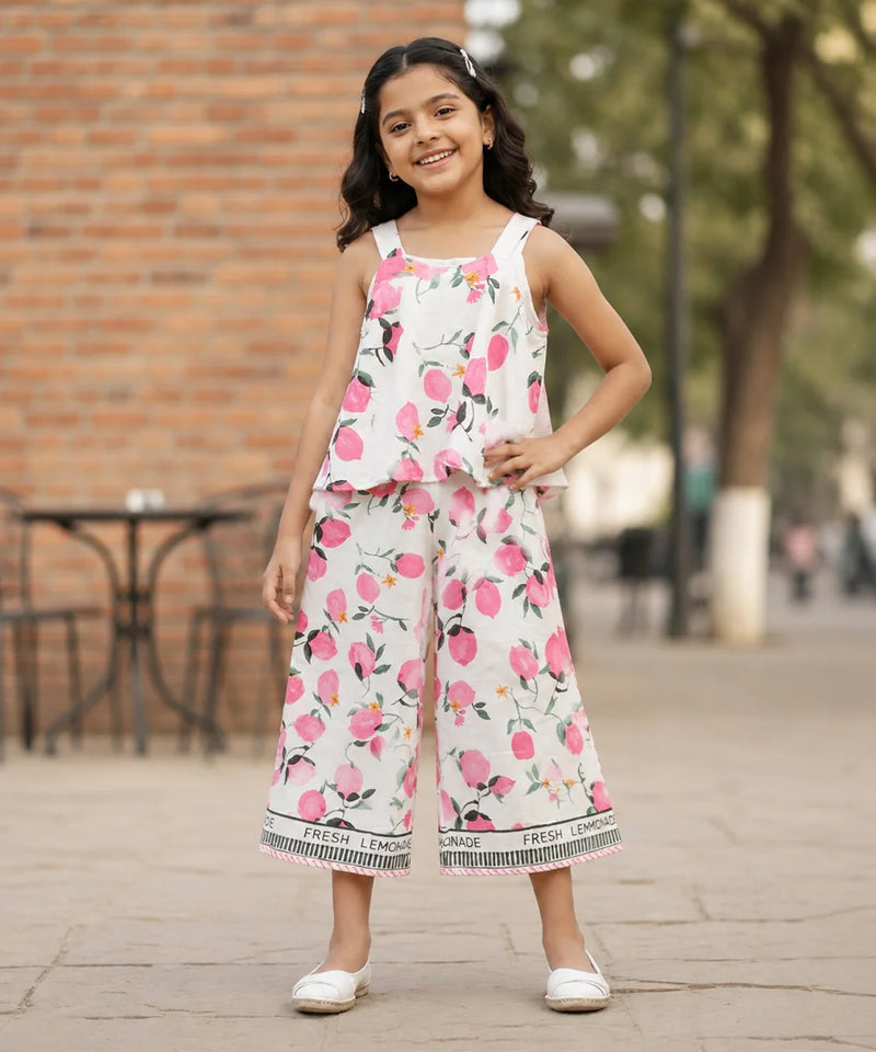 A young girl is shown from behind wearing a white sleeveless jumpsuit with a pink and green watercolor floral print. The jumpsuit features ruffled straps and a wide leg, with a black and white patterned hem that reads 'LEMONADE'.
