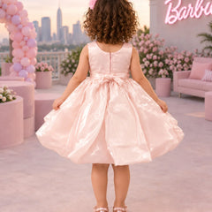 A young girl stands outdoors wearing a shiny light pink party dress with a large gray and pink flower applique at the chest. The background shows a pink-themed patio with balloons, flowers, a sofa, and a neon sign.