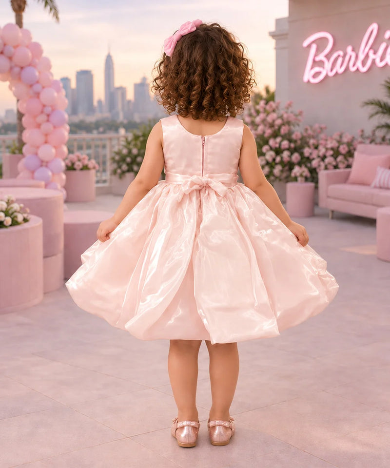 A young girl stands outdoors wearing a shiny light pink party dress with a large gray and pink flower applique at the chest. The background shows a pink-themed patio with balloons, flowers, a sofa, and a neon sign.