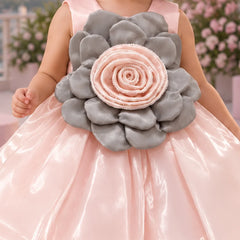 A young girl stands outdoors wearing a shiny light pink party dress with a large gray and pink flower applique at the chest. The background shows a pink-themed patio with balloons, flowers, a sofa, and a neon sign.