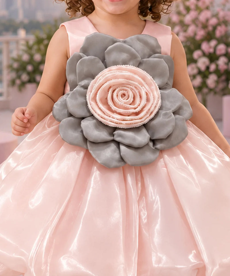 A young girl stands outdoors wearing a shiny light pink party dress with a large gray and pink flower applique at the chest. The background shows a pink-themed patio with balloons, flowers, a sofa, and a neon sign.