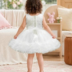 A toddler girl viewed from behind wearing a sleeveless white tulle flower girl dress with a beaded bodice and a ruffled skirt, standing in a softly decorated room.