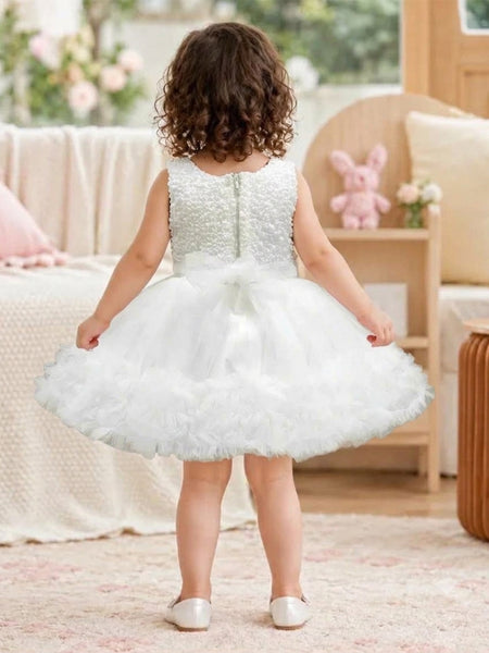 A toddler girl viewed from behind wearing a sleeveless white tulle flower girl dress with a beaded bodice and a ruffled skirt, standing in a softly decorated room.