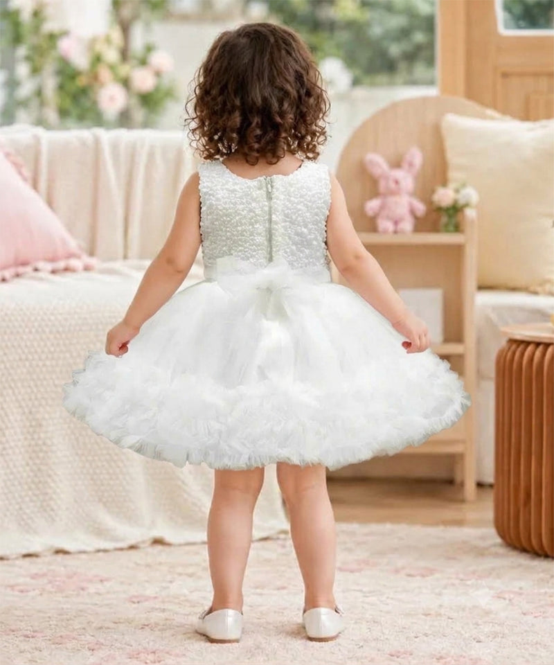 A toddler girl viewed from behind wearing a sleeveless white tulle flower girl dress with a beaded bodice and a ruffled skirt, standing in a softly decorated room.