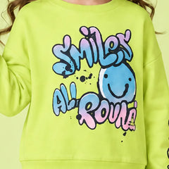 A close-up of a child wearing a bright lime green sweatshirt with a colorful graphic that reads 'SMILEY ALL ROUND' in blue, pink, and purple bubble letters, alongside a smiling blue face.