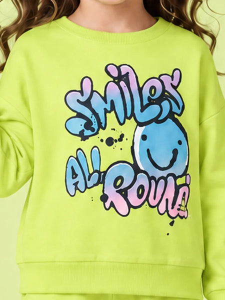 A close-up of a child wearing a bright lime green sweatshirt with a colorful graphic that reads 'SMILEY ALL ROUND' in blue, pink, and purple bubble letters, alongside a smiling blue face.