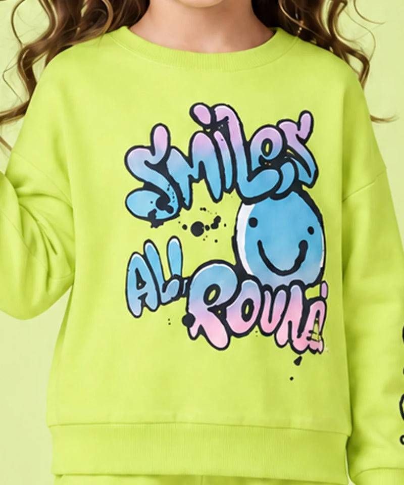 A close-up of a child wearing a bright lime green sweatshirt with a colorful graphic that reads 'SMILEY ALL ROUND' in blue, pink, and purple bubble letters, alongside a smiling blue face.