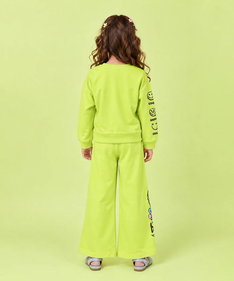 A child model shown from the back wearing a bright lime green two-piece tracksuit with a sweatshirt and wide-leg pants, featuring black graphic text on the sleeves and leg, set against a solid lime green background.
