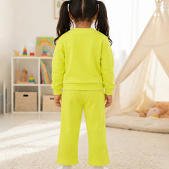 A young girl with dark hair in pigtails stands with her back to the camera wearing a bright neon yellow-green two-piece lounge set consisting of a long-sleeve sweatshirt and matching wide-leg pants, in a brightly lit children's room.