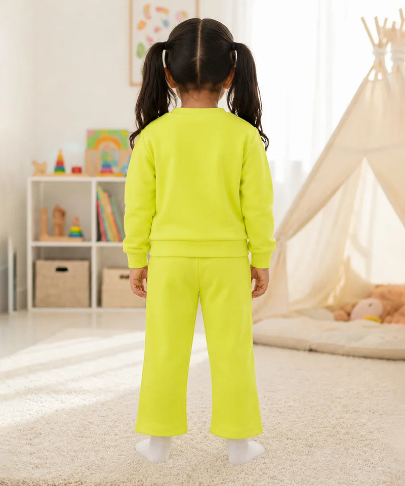A young girl with dark hair in pigtails stands with her back to the camera wearing a bright neon yellow-green two-piece lounge set consisting of a long-sleeve sweatshirt and matching wide-leg pants, in a brightly lit children's room.