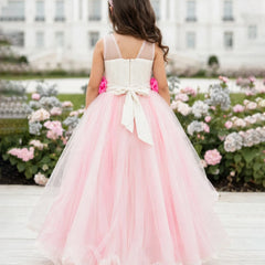 A young girl viewed from behind wearing a formal pink and white tulle flower girl dress with a large bow at the back, standing outdoors in front of a white building and flower beds.
