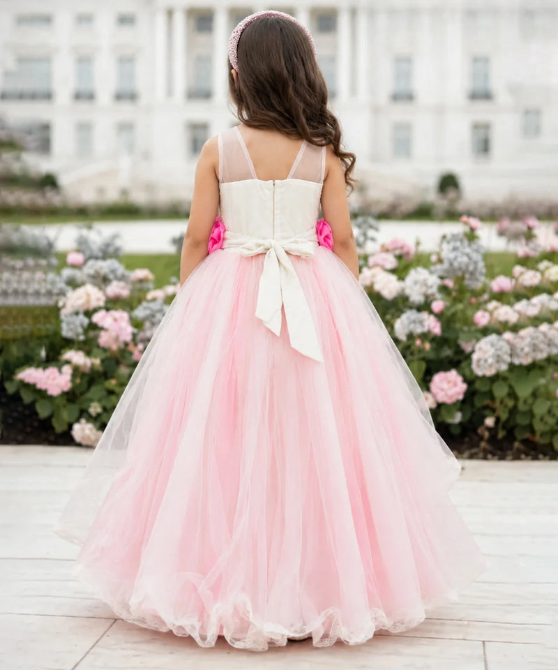 A young girl viewed from behind wearing a formal pink and white tulle flower girl dress with a large bow at the back, standing outdoors in front of a white building and flower beds.