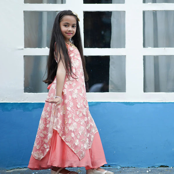 Young girl in a pink floral dress standing in front of a white window frame with blue wall.