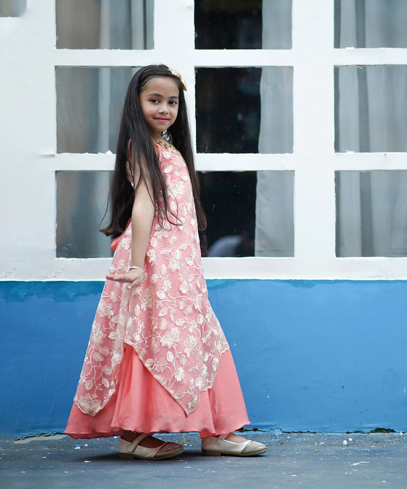 Young girl in a pink floral dress standing in front of a white window frame with blue wall.