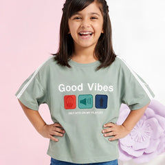 A young girl with dark hair smiles while wearing a sage green graphic t-shirt with white sleeve stripes. The shirt features the text 'Good Vibes' and 'ONLY HITS ON MY PLAYLIST' above three colored icons (red headphones, teal megaphone, blue arrow).