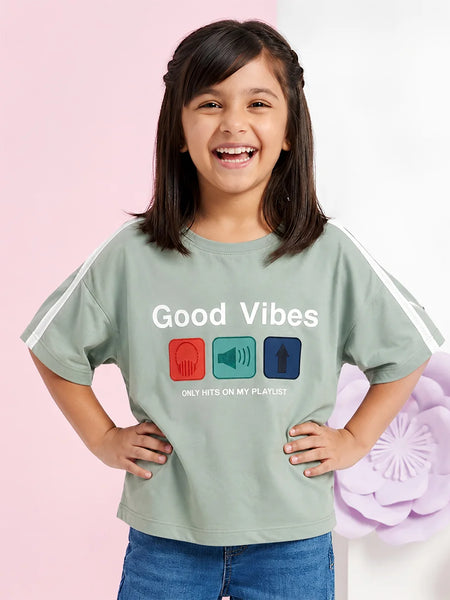 A young girl with dark hair smiles while wearing a sage green graphic t-shirt with white sleeve stripes. The shirt features the text 'Good Vibes' and 'ONLY HITS ON MY PLAYLIST' above three colored icons (red headphones, teal megaphone, blue arrow).