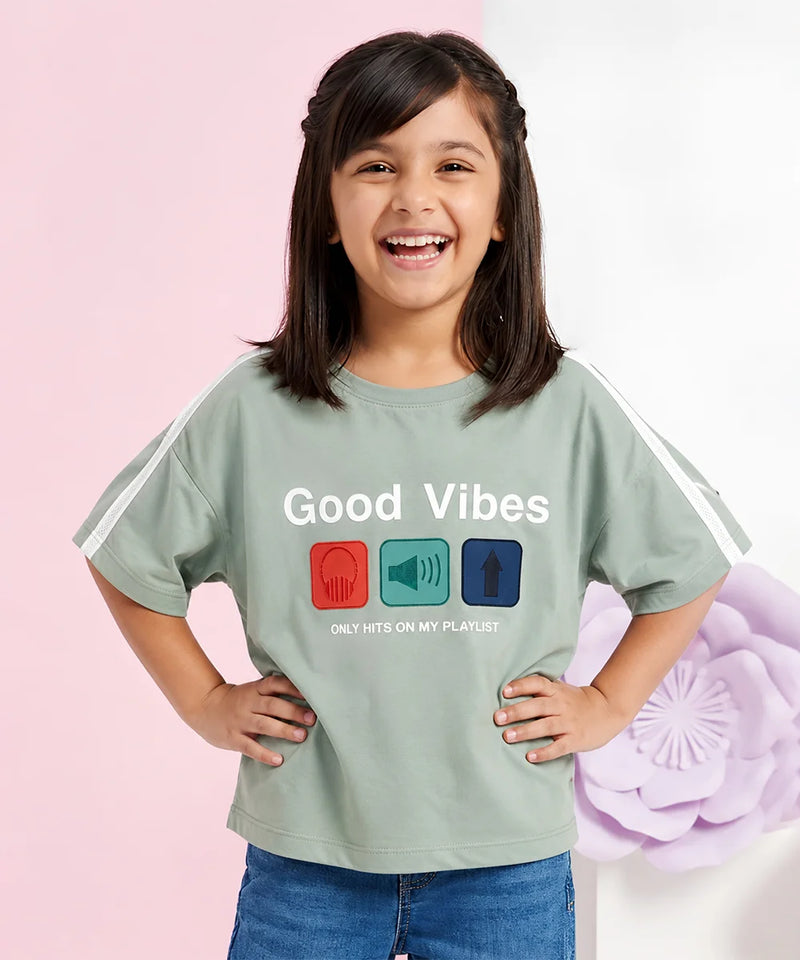 A young girl with dark hair smiles while wearing a sage green graphic t-shirt with white sleeve stripes. The shirt features the text 'Good Vibes' and 'ONLY HITS ON MY PLAYLIST' above three colored icons (red headphones, teal megaphone, blue arrow).