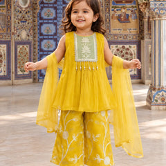 A young girl stands in a decorated courtyard wearing a bright yellow traditional outfit with a light green embroidered bodice panel, matching wide-leg pants with white floral embroidery, and sheer yellow dupatta sleeves.