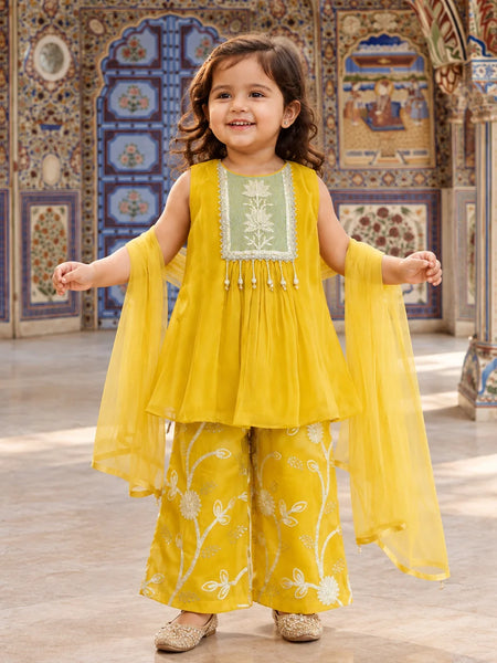 A young girl stands in a decorated courtyard wearing a bright yellow traditional outfit with a light green embroidered bodice panel, matching wide-leg pants with white floral embroidery, and sheer yellow dupatta sleeves.