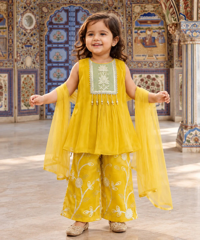 A young girl stands in a decorated courtyard wearing a bright yellow traditional outfit with a light green embroidered bodice panel, matching wide-leg pants with white floral embroidery, and sheer yellow dupatta sleeves.