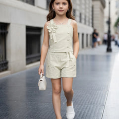 A young girl walking down a city street wearing a light beige linen-look sleeveless romper with decorative buttons, a matching fabric flower applique on the chest, and a tie belt at the waist. She is carrying a small white handbag and wearing white slip-on shoes.