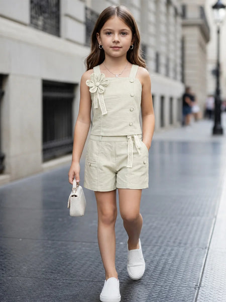 A young girl walking down a city street wearing a light beige linen-look sleeveless romper with decorative buttons, a matching fabric flower applique on the chest, and a tie belt at the waist. She is carrying a small white handbag and wearing white slip-on shoes.