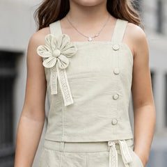 A child wearing a light beige linen-look sleeveless top with wide straps, decorative buttons, and a large fabric flower applique on the chest, paired with matching shorts.
