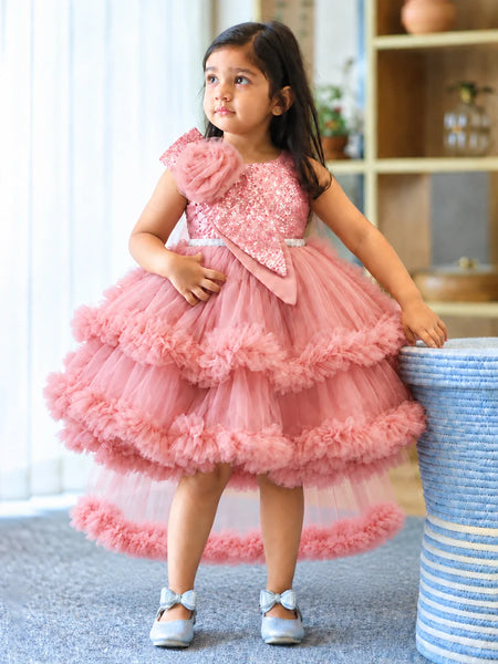 A young girl wearing a pink tiered tulle party dress with a sequined bodice and ruffled layers, standing indoors next to a blue-and-white striped basket.