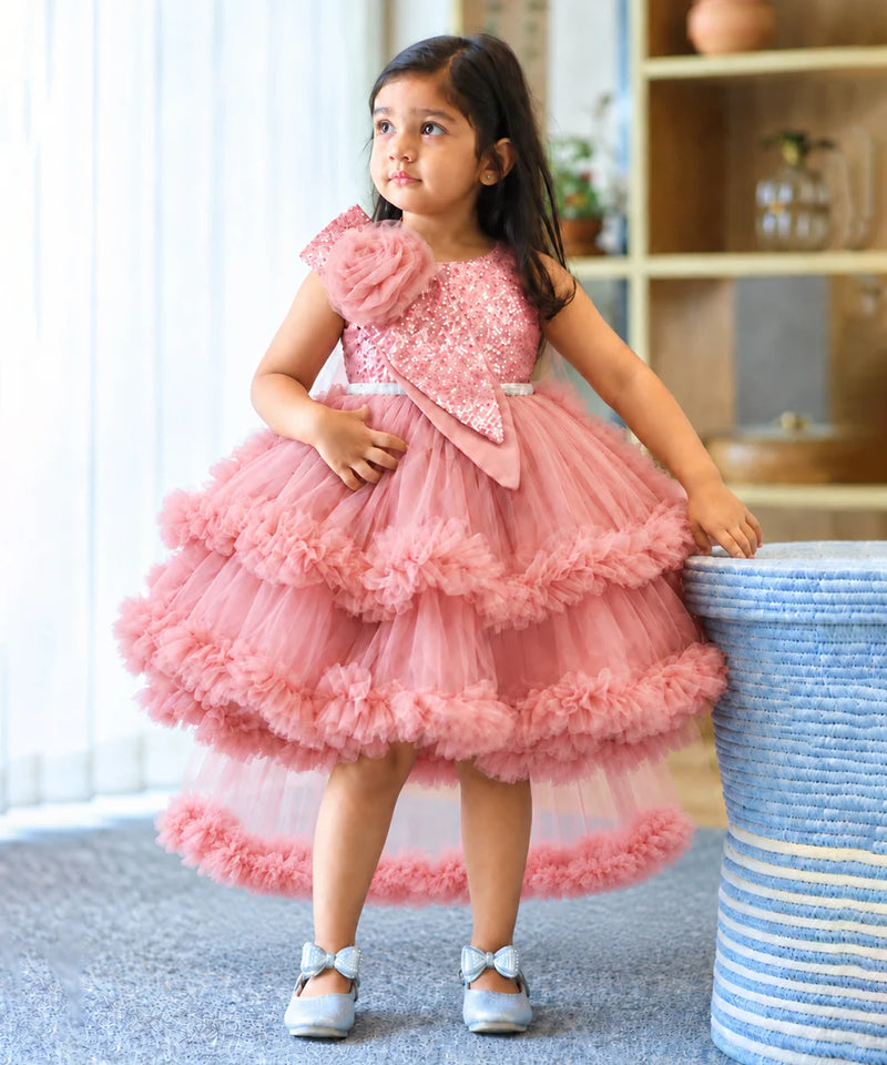 A young girl wearing a pink tiered tulle party dress with a sequined bodice and ruffled layers, standing indoors next to a blue-and-white striped basket.