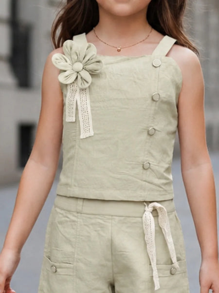 A child wearing a sleeveless light sage green linen romper with a large fabric flower applique on the shoulder, decorative buttons down the front, and matching shorts with a drawstring waist.