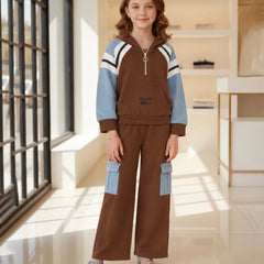 A young girl stands in a bright, modern indoor room wearing a two-piece tracksuit. The set features a brown pullover hoodie with light blue and white color-blocked sleeves, and matching brown wide-leg pants with light blue cargo pockets.