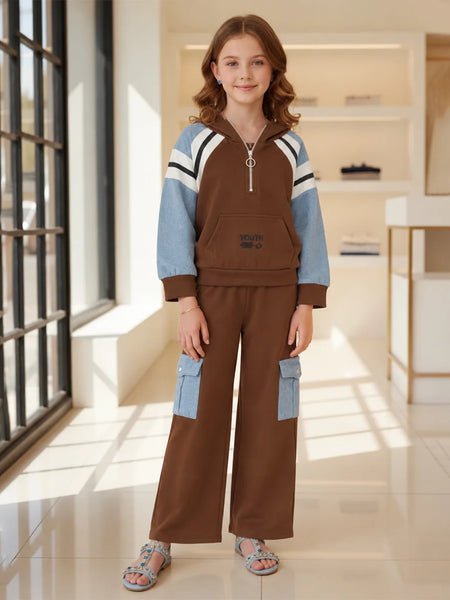 A young girl stands in a bright, modern indoor room wearing a two-piece tracksuit. The set features a brown pullover hoodie with light blue and white color-blocked sleeves, and matching brown wide-leg pants with light blue cargo pockets.