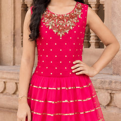 A young girl wearing a bright pink sleeveless dress with gold polka dots, a heavily embroidered neckline, and a flared tulle skirt with gold horizontal stripes, posed in front of a stone architectural background.