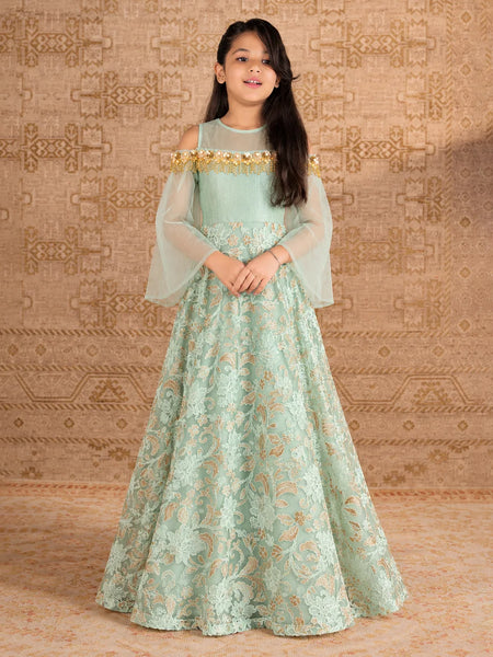 A woman viewed from behind wearing a mint green off-the-shoulder gown with gold floral embroidery, sheer sleeves, and a matching waist tie, set against a patterned beige background.