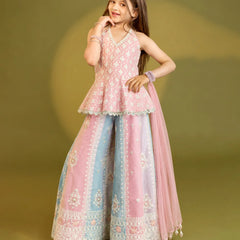 A young girl modeling a traditional Indian outfit consisting of a light pink embroidered sleeveless top, wide-leg pink and light blue pants with white embroidery, and a matching sheer pink dupatta, set against a solid olive green studio background.