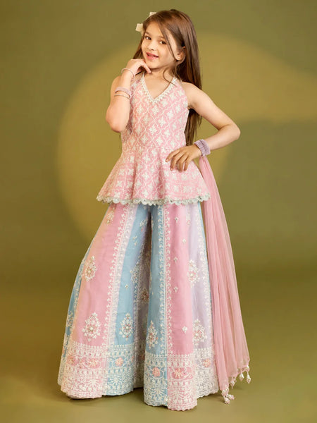 A young girl modeling a traditional Indian outfit consisting of a light pink embroidered sleeveless top, wide-leg pink and light blue pants with white embroidery, and a matching sheer pink dupatta, set against a solid olive green studio background.